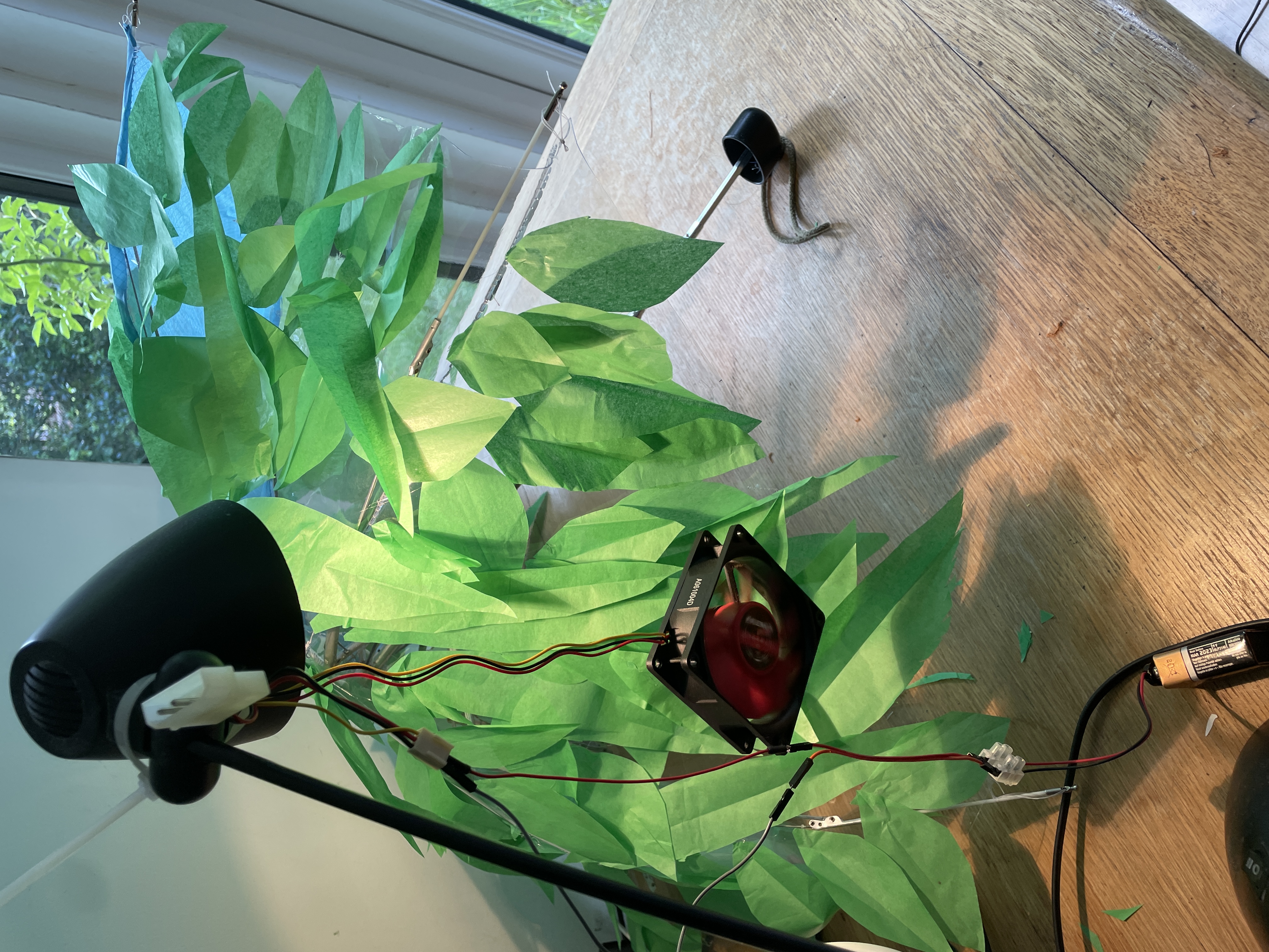 A fan cable-tied to a lamp, which is shining on an umbrella on a desk. the umbrella has a load of paper leaves stuck to it.