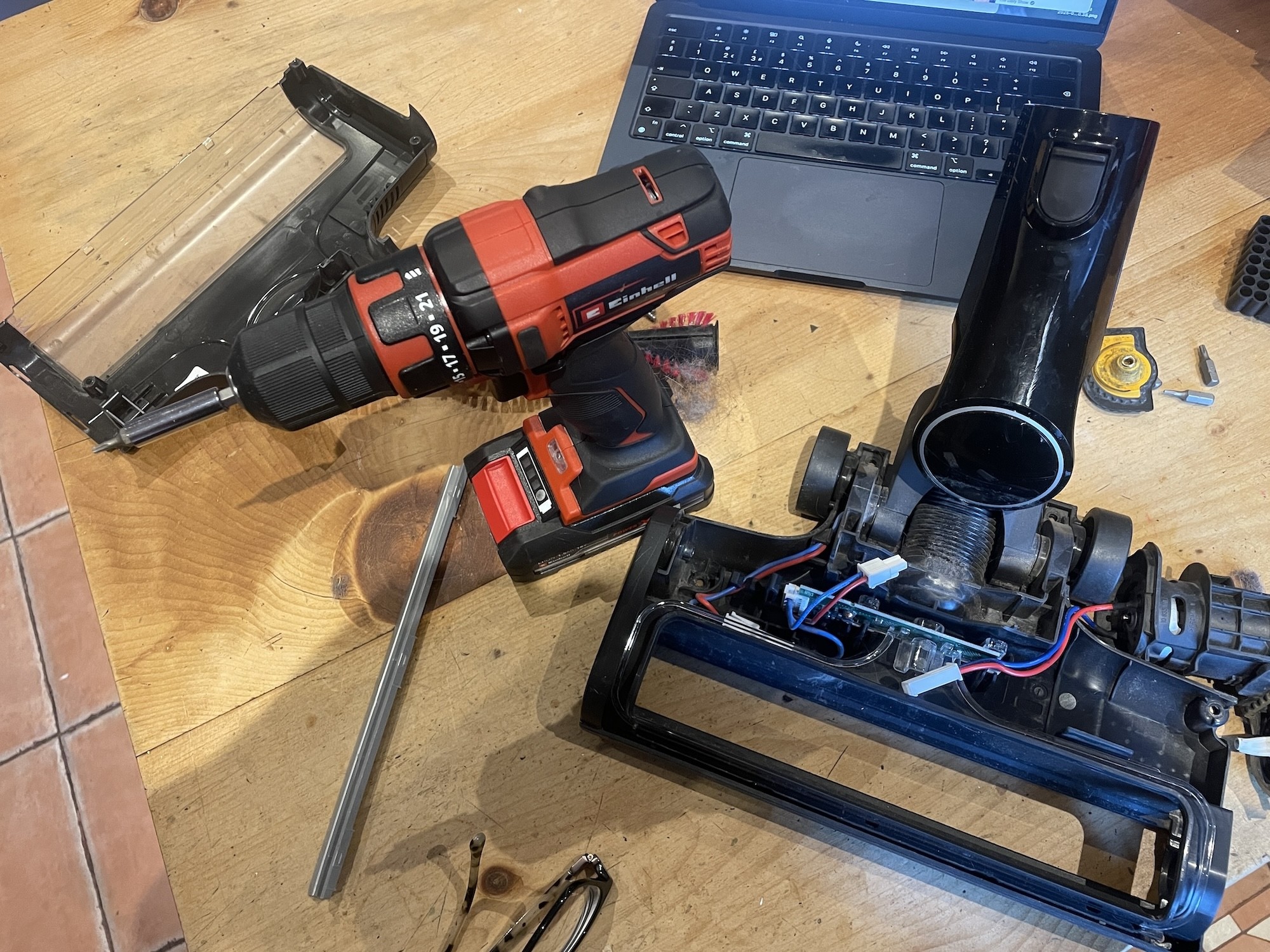 a vaccum cleaner head completely taken apart on a wooden table acommpanied by an electric drill and a laptop